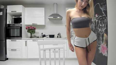 Media: Video of a blonde, fair-skinned woman in a tight, grey crop top and white shorts, posing in a minimalist, white kitchen with black appliances and a chalkboard wall.