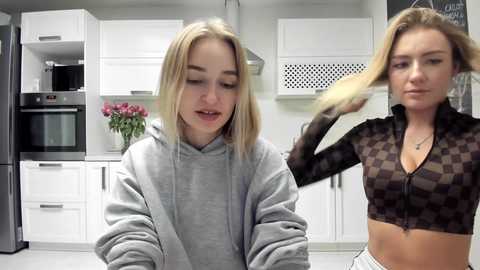 Media: Video of two young women in a modern kitchen; one blonde in a grey hoodie, the other blonde with a patterned crop top.