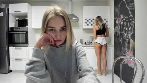 Media: Video of a blonde woman in a grey hoodie, leaning on a kitchen table, while a slim, tan-skinned woman in a black top and white shorts cooks in the background.