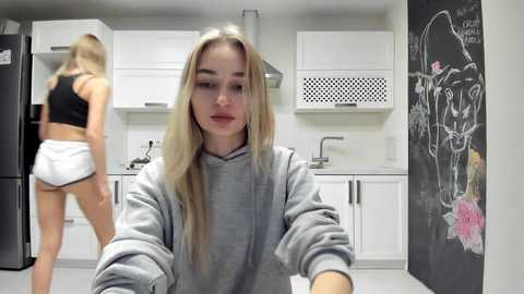 Media: Video of a blonde woman in a gray hoodie and white shorts, standing in a modern kitchen with white cabinets, a blackboard, and a blurred background.