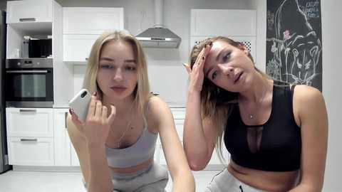 Media: Video of two young Caucasian women in modern kitchen. One blonde in white tank top, other brunette in black sports bra, both looking tired, holding heads.