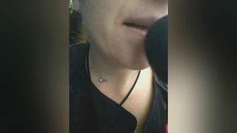 Video of a woman's face and neck, wearing a black shirt with a thin strap, partially obscured by a large microphone. Background is blurred with greenery.
