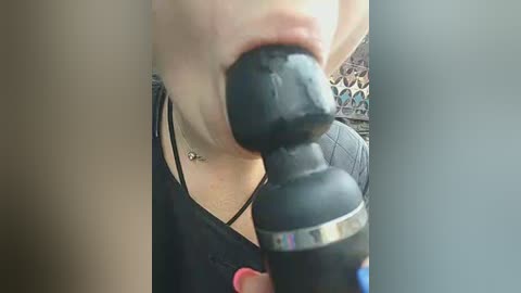 Video of a woman in a black dress with a necklace, using a black vibrator on her face. Background shows a mosaic pattern.