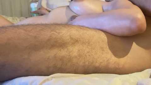 Video of a close-up view of a man's hairy, muscular thigh and partially visible genitalia, with a woman's bare breasts and nipples visible in the background, lying on a white bedsheet in a dimly lit room.