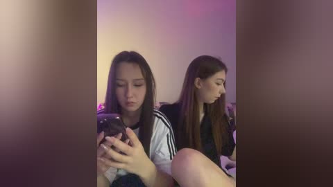 Media: A video of two young women with long hair, one holding a phone, the other in the background, against a dimly lit, purple-hued wall.