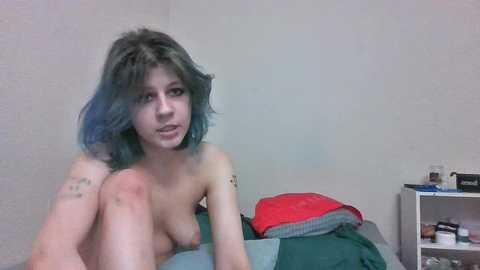 Media: Video of a young, topless woman with shoulder-length, blue-dyed hair, sitting on a bed with messy bedding, against a plain, light-colored wall. She has a tattoo on her right arm.