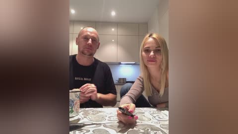 Media: A video of a Caucasian couple, a man with short hair in a black t-shirt and a woman with long blonde hair in a gray sweater, eating at a table with a patterned tablecloth.