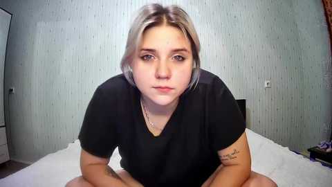 Media: Video of a young, fair-skinned, blonde-haired woman with tattoos on her arms, wearing a black t-shirt, sitting on a bed with white sheets in a simple, gray wallpapered room.