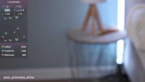 Video of a dimly lit bedroom with a small, light-colored bedside table featuring a white lamp and a brown wooden tripod. The background shows a blurry, light blue wall. The image is soft-focused with a watermark \"yourprincessalina\" at the bottom.
