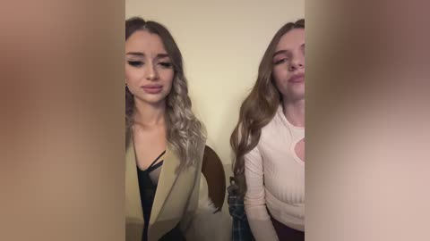 Media: Video of two young women with long hair and different clothing styles standing side-by-side in front of a beige wall. One wears a beige blazer over a black top, while the other sports a white long-sleeve top.