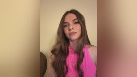 Media: Video of a young woman with long, wavy brown hair, wearing a bright pink sleeveless top, standing in a beige-walled room with a circular mirror to her left.