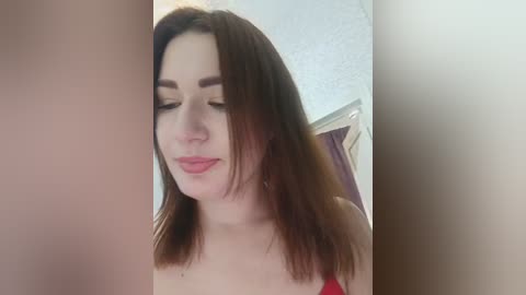 Media: Video of a young woman with fair skin and shoulder-length auburn hair, wearing a red top, with a neutral expression, in a softly lit room.