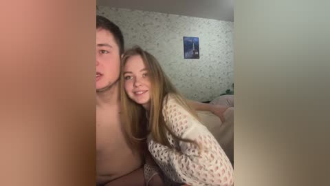 Video of a young couple, a shirtless man with short hair and a smiling woman with long brown hair, in a cozy bedroom with floral wallpaper and a blue poster.
