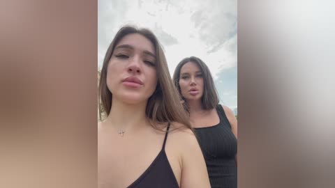 Media: Video of two young women with fair skin and straight, shoulder-length brown hair, wearing black tank tops. One woman has a slight pout, while the other has a neutral expression. The background features a cloudy sky.