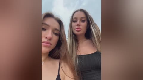 Media: Video of two young women with long hair, one in a black top, the other in a grey top, standing close together against a cloudy sky background.