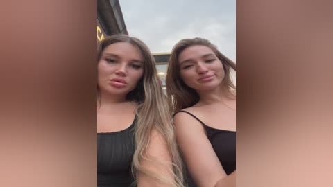 Media: Video of two young women, both with long, straight brown hair, wearing black tank tops, standing close together outdoors with a cloudy sky in the background.