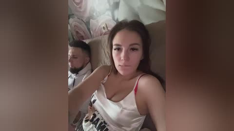 Media: Video of a young woman with long brown hair, wearing a white camisole, sitting on a bed with a man in the background, both looking into a mirror.
