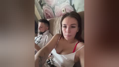 Media: Video of a young woman with long brown hair, wearing a white tank top, seated next to a bearded man in a hoodie, both on a beige couch.