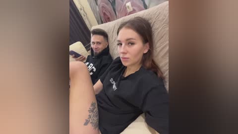 A video of a young couple sitting on a beige couch, both wearing black t-shirts. The woman, with long brown hair, holds a book, revealing a tattoo on her thigh. The man, with short dark hair, appears relaxed.