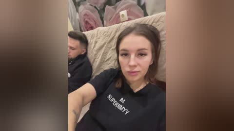 Media: Video of a young woman with light skin and straight brown hair, wearing a black t-shirt with white text, seated on a beige couch with floral pillows in the background.