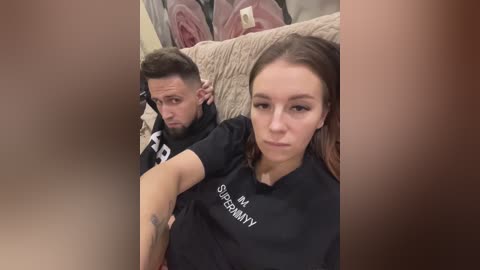 Media: A video of a couple sitting on a couch, the man with short dark hair and beard, the woman with long brown hair, both wearing black t-shirts. Background includes beige couch and pink blanket.