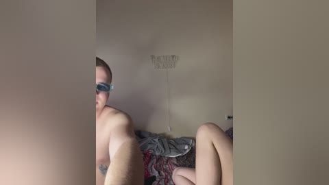Video of a shirtless, muscular man with a tattoo on his left shoulder, wearing sunglasses, lying on a bed with red and black patterned sheets, in a dimly lit room.