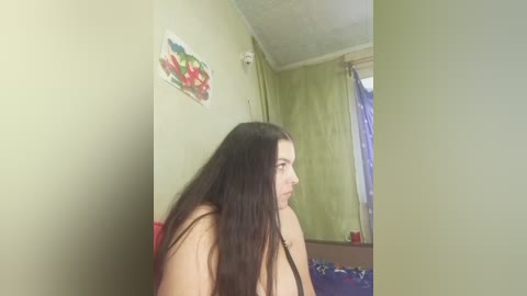 Video of a young woman with long dark hair, bare chest, and fair skin, sitting indoors, wearing a black top, in a room with green walls and a colorful floral painting.