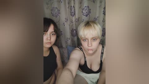 Media: Video of two women with short hair, one with dark hair and the other blonde, wearing tank tops, sitting on a bed with floral curtains in the background.