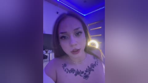 Media: Video of a young woman with fair skin, dark hair, and a floral tattoo on her chest, standing in a dimly lit room with blue LED lighting.