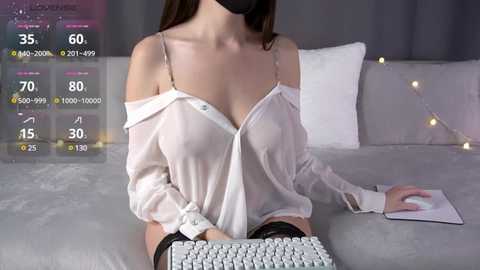 Video of a fair-skinned woman in a sheer white blouse, revealing cleavage, black mask, and sitting on a bed with a keyboard.