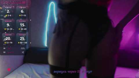 Video of a person wearing black lingerie and a mask, standing in a dimly lit room with neon lights and a bed in the background.