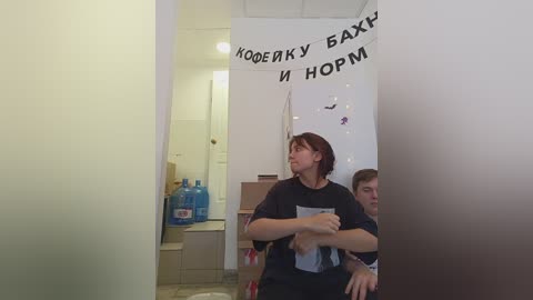 Media: Video of a woman with shoulder-length brown hair in a dark t-shirt, standing in a modern, sparsely furnished room with beige walls and a white door. She holds a whiteboard with Russian text.