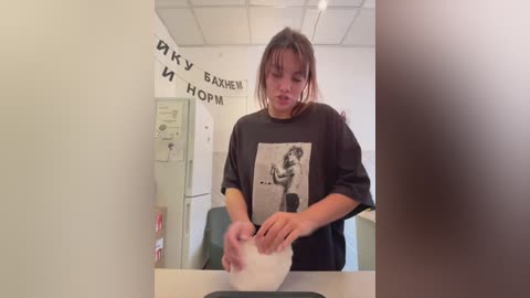 Media: Video of a young woman with shoulder-length brown hair in a plain white room, wearing a black t-shirt with a photo of a woman in a bathtub. She is pouring liquid from a pitcher into a sink.