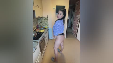 Media: Video of a young woman with long dark hair, wearing a blue and white checkered shirt and black thong, standing in a modern kitchen with white cabinets, a brick wall, and a tiled backsplash.