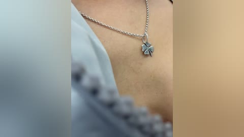 Media: A close-up video of a person's upper torso, wearing a silver necklace with a clover charm. The background is blurred, with a focus on the necklace and skin texture. The individual is wearing a light-colored, possibly blue, garment.