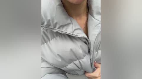Media: A video of a person wearing a light gray, puffy winter jacket with a high collar, zipped up to their chin. The background is a plain, light gray wall.