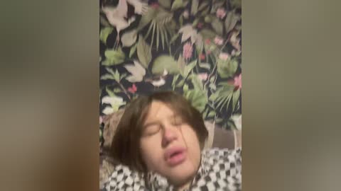 Video of a young person with light skin and shoulder-length brown hair, eyes closed in apparent ecstasy, set against a vibrant floral wallpaper backdrop.