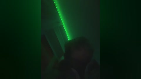 A dimly lit video shows a person with short hair, blurry and indistinct, in front of a glowing green neon light. The background is dark, creating a moody, mysterious atmosphere.