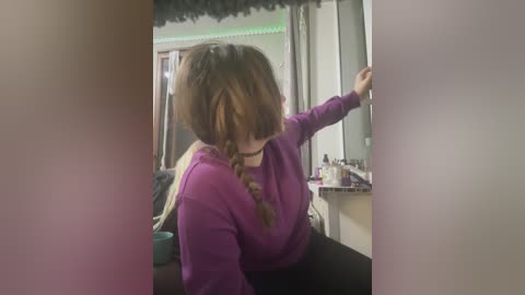 Video of a young woman with long brown hair in braids, wearing a purple sweater, leaning against a mirror in a dimly lit, cluttered bathroom.