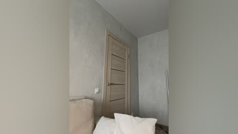 Media: Video of a minimalist, sparsely furnished bedroom with light gray walls, a wooden door, a beige cushioned headboard, and white bedding.