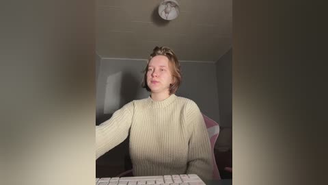 Media: Video of a young Caucasian woman with light brown hair, wearing a cream-colored sweater, sitting at a desk with a white keyboard, in a dimly lit room with gray walls and a ceiling light.