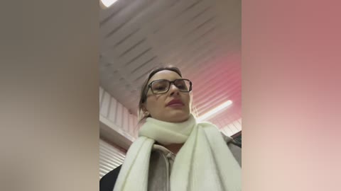 A video of a woman with light skin, dark hair, and glasses, wearing a green scarf, standing in an indoor space with a white ceiling and pink lighting.