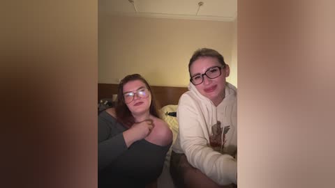 A video captures two women with glasses, one in a gray off-shoulder top, the other in a white hoodie, seated in a dimly-lit room with beige walls and a wooden headboard.