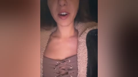 Media: Video of a woman with light skin and brown hair, wearing a beige lace-up corset top and a beige cardigan, partially visible. She has a slightly open mouth, possibly in mid-speech.