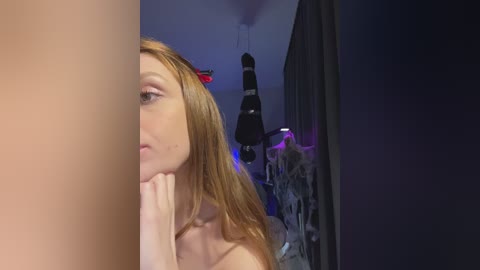 Video of a pale-skinned, red-haired woman with a small red devil horn headband, nude, in a dimly lit room with dark curtains and a hanging light fixture.