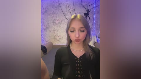 Media: Video of a young woman with long, straight blonde hair, wearing a black corset top with pearl accents, standing in front of a floral wallpaper with a spider web and blue lights, looking down.