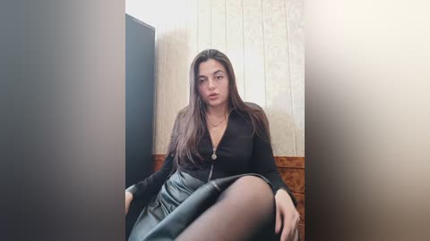 Media: Video of a young woman with long brown hair, wearing a black zip-up top and black leather skirt, seated indoors with beige wallpaper and wooden paneling background.