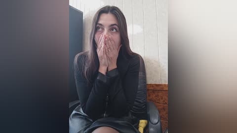 Media: Video of a woman with long dark hair, wearing a black jacket, seated on a chair, hands covering her mouth in surprise. Background shows a white tiled wall and wooden furniture.