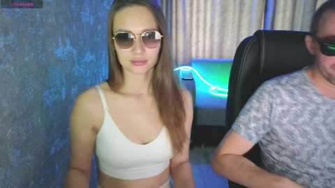 Video of a young Caucasian woman with long brown hair, wearing a white crop top, aviator sunglasses, and sitting beside a man in a gray shirt. Background features a blue wall and a lit-up gaming setup.