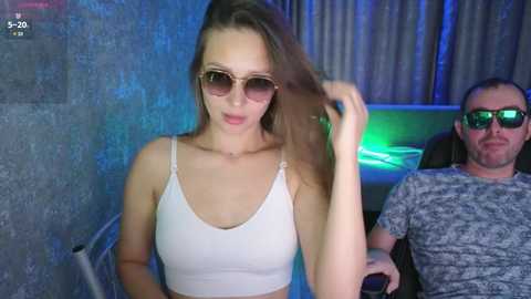 Video of a young Caucasian woman with light skin, brown hair, and sunglasses, wearing a white crop top, standing beside a bald man in a grey shirt, both smiling in a dimly lit, blue-lit room.